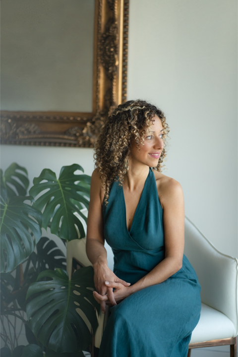 Bailey Vaez smiling, seated in a teal dress near a plant in a softly lit room